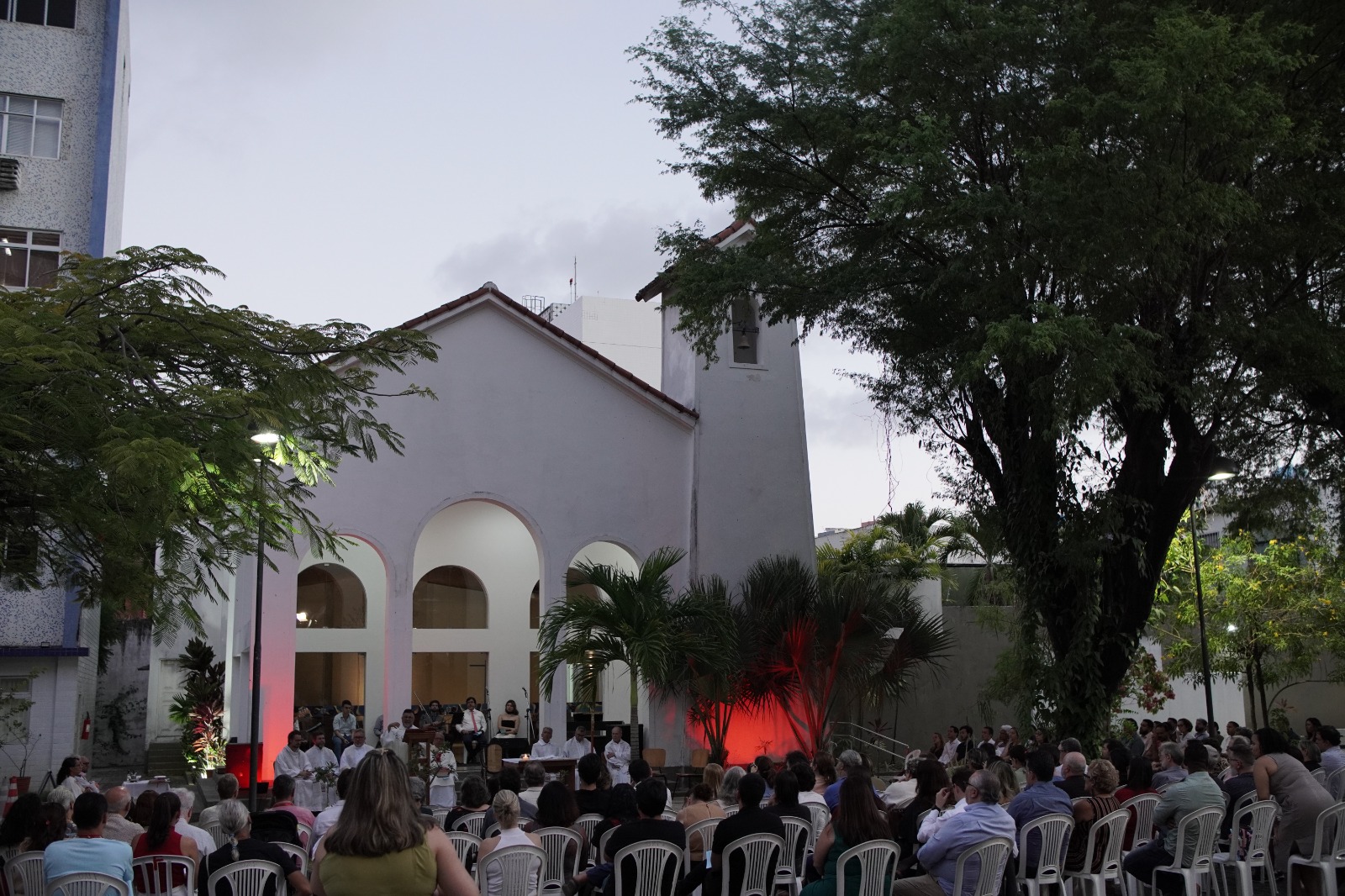 A imagem retrata a celebração da Missa de Natal ao ar livre, no adro da Capela Santo Inácio de Loyola e Companheiros Jesuítas, localizada no campus Recife da Universidade Católica de Pernambuco (Unicap). A capela, de fachada branca simples e elegante, com telhado de telhas vermelhas inclinadas, uma torre sineira quadrada à direita com abertura para sino no topo e três grandes arcos ogivais na entrada principal iluminados internamente por luz suave, serve como pano de fundo para o evento. No centro da cena, nos degraus da entrada da capela transformados em altar improvisado, um grupo de cerca de 10 a 15 pessoas participa da celebração: padres ou celebrantes de vestes litúrgicas brancas, possivelmente com um presbítero principal ao centro, acompanhados por leitores, coroinhas ou ministros, microfones, um teclado portátil e equipamentos de som, sugerindo momentos de cantos natalinos ou leituras. Em primeiro plano, uma numerosa assembleia de aproximadamente 100 a 150 fiéis está sentada em fileiras de cadeiras plásticas brancas arrumadas em semicírculo ou linhas paralelas, voltadas para a capela. O público é composto por adultos, idosos e famílias, vestindo roupas leves e casuais adequadas ao clima tropical, muitos de perfil ou de costas, atentos e participativos à missa. O ambiente é o pátio arborizado do campus, enquadrado por grandes árvores de folhagem densa e verde escura à esquerda e à direita, palmeiras e plantas tropicais no centro, uma delas destacada por uma iluminação vermelha intensa vinda de baixo, criando um efeito dramático e festivo nas folhas, possivelmente simbolizando o espírito natalino. Postes de luz externa estão acesos, indicando que a celebração ocorre ao entardecer ou início da noite, sob um céu nublado e cinzento. A atmosfera transmite serenidade, comunhão e alegria comunitária típica de uma Missa de Natal universitária jesuíta, com contraste entre o branco puro da capela e das cadeiras, o verde exuberante das árvores e o toque vermelho da iluminação especial, em um espaço acolhedor e arborizado no coração do Recife.