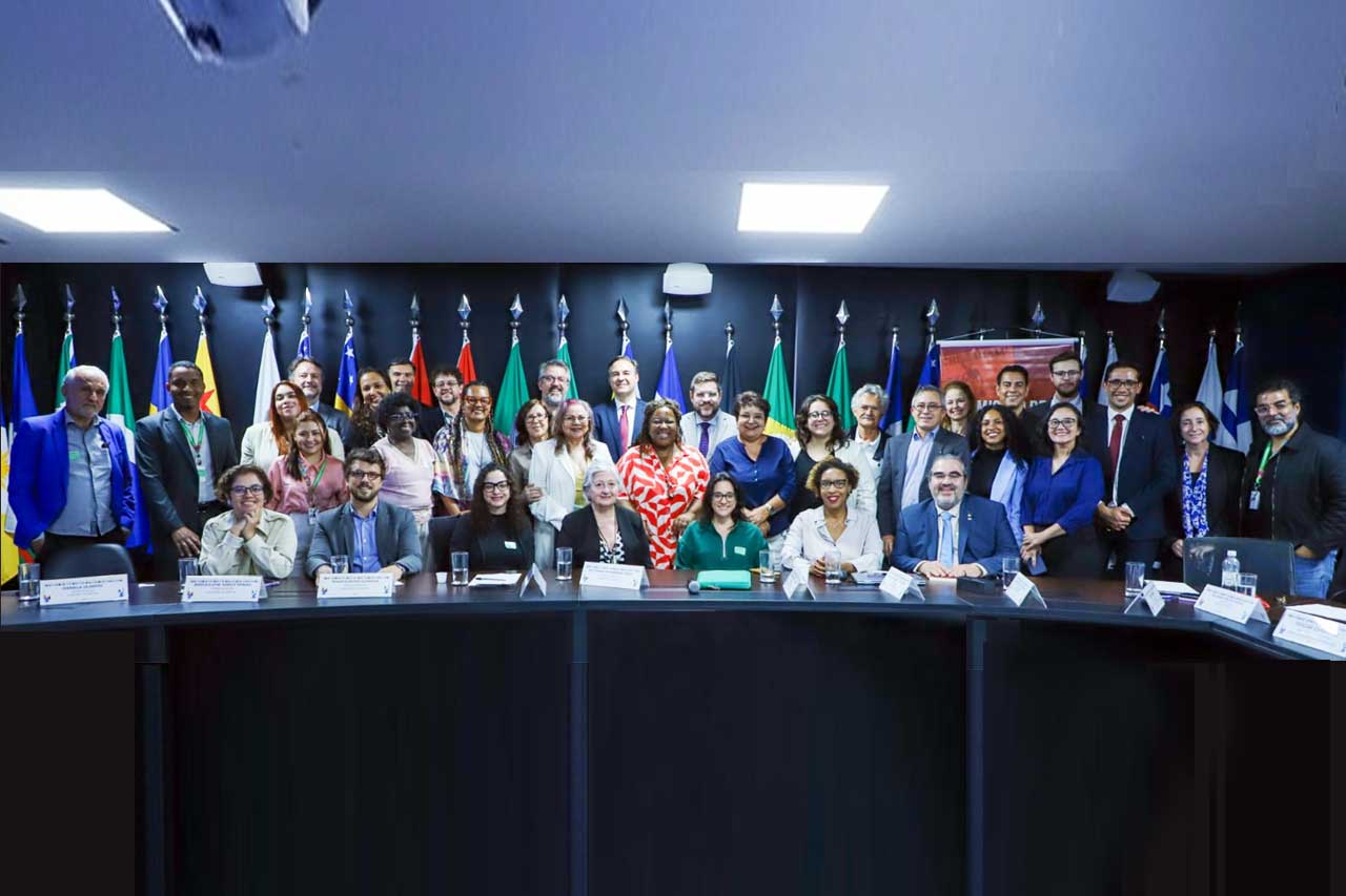 Fotografia de um grupo grande, cerca de 30 pessoas, posando para uma foto oficial em um auditório ou sala de reuniões. As pessoas estão organizadas em três fileiras: A primeira fileira está sentada à mesa de reuniões semicircular, com placas de identificação à frente de cada um; A segunda fileira está em pé logo atrás dos que estão sentados; A terceira fileira está mais ao fundo, em pé. Ao fundo, há uma parede preta com várias bandeiras dos estados do Brasil penduradas em mastros, dispostas em linha. À direita, há um banner vermelho com texto parcialmente visível.A maioria das pessoas usa roupas formais ou semiformales (camisas, blazers, vestidos), muitas com crachás de identificação pendurados no pescoço. O clima parece alegre: quase todos sorriem ou fazem expressões amigáveis para a câmera. A iluminação é clara e uniforme, vinda do teto.A composição transmite a ideia de um evento institucional, provavelmente uma reunião, seminário ou comissão com representantes de diferentes estados brasileiros.