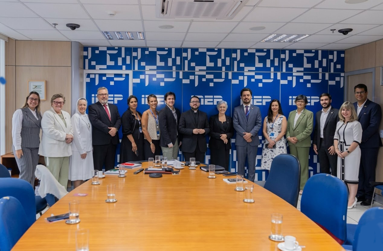 A imagem mostra um grupo de 15 pessoas posando para uma foto oficial em uma sala de reuniões formal. Elas estão alinhadas horizontalmente ao fundo da sala, atrás de uma grande mesa de madeira clara que domina o primeiro plano. Ao fundo, há um painel azul vibrante com logotipos repetidos e grafismos em branco.