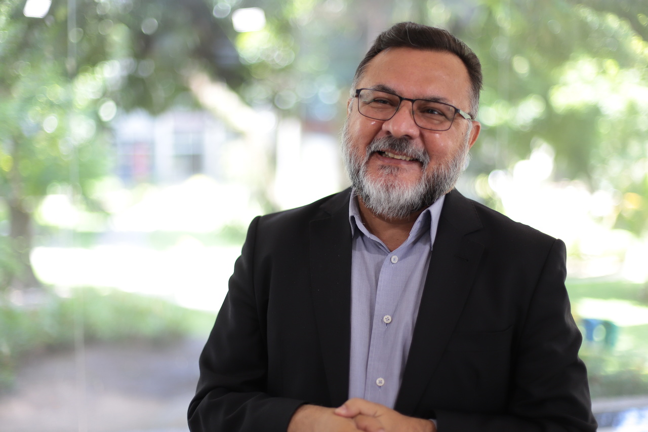 Padre Pedro sorrindo em foto de plano americano no salão de vidro