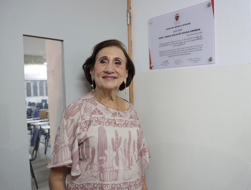 Zélia em frente à placa no auditório que leva seu nome